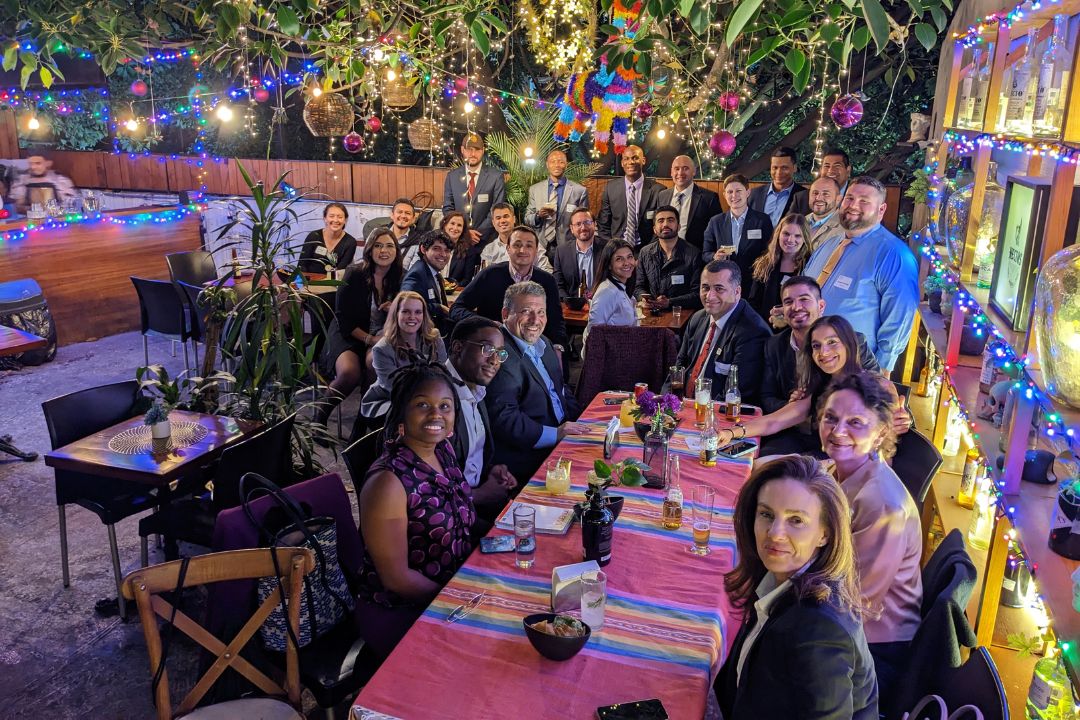 Large group of alumni smiling at dinner table in a restaurant garden decorated with brightly lit and colorful string lights
