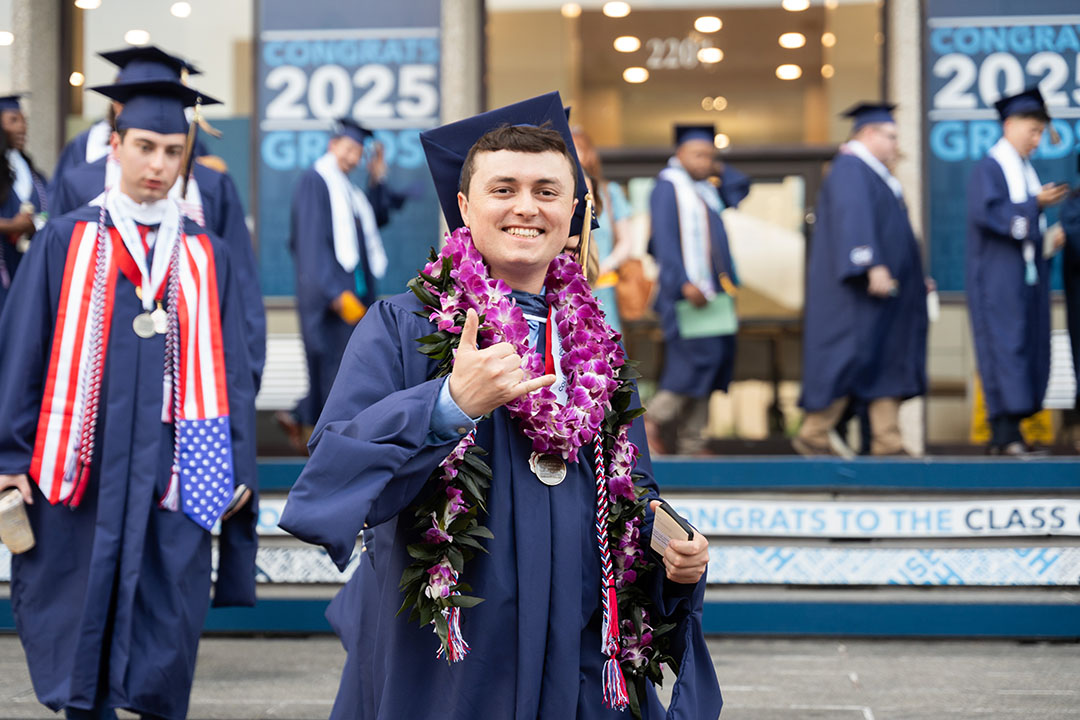 Man in commencement gown and cap with thumbs up, other graduates and 2025 signs in the background